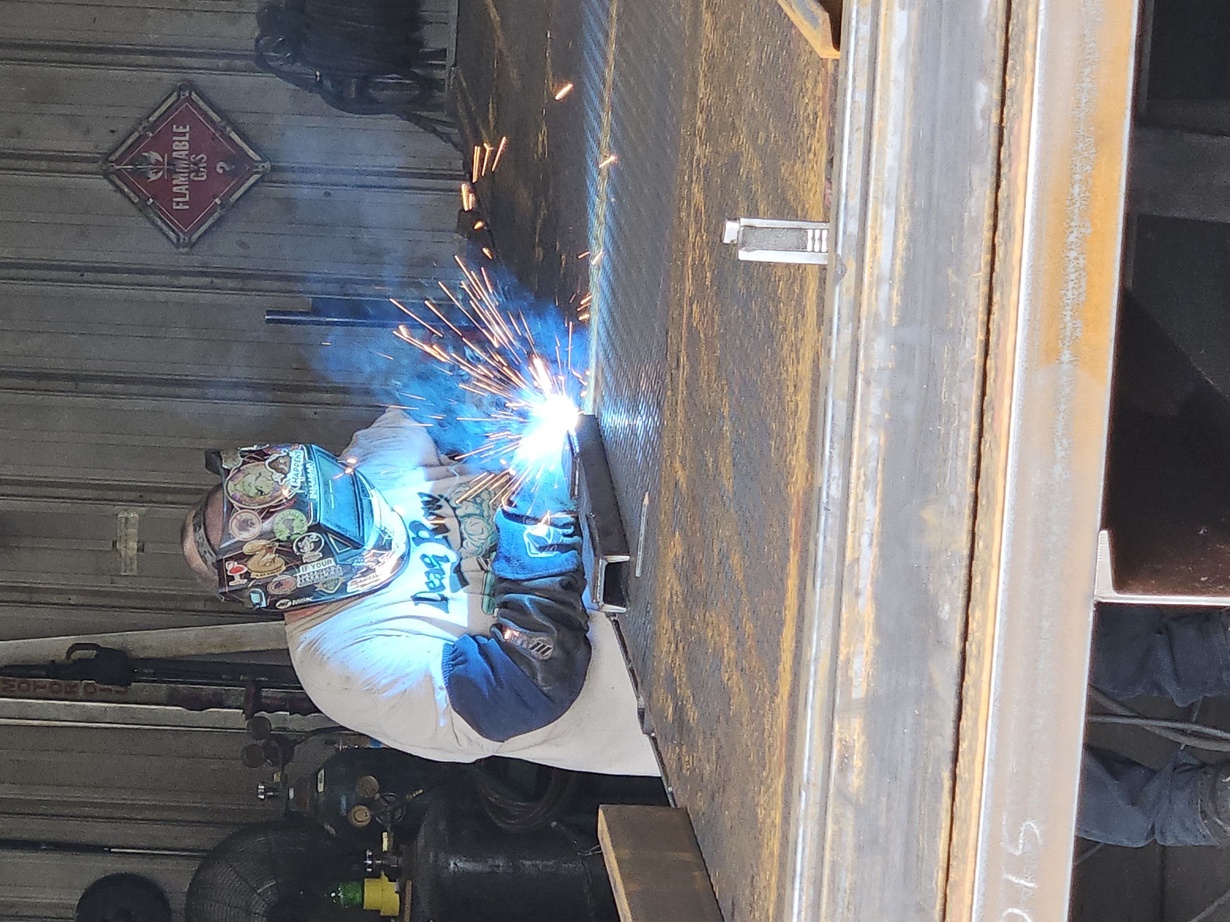 Welder in a metal fabrication shop using a torch to weld steel on a large flatbed surface, with bright sparks and protective gear visible.